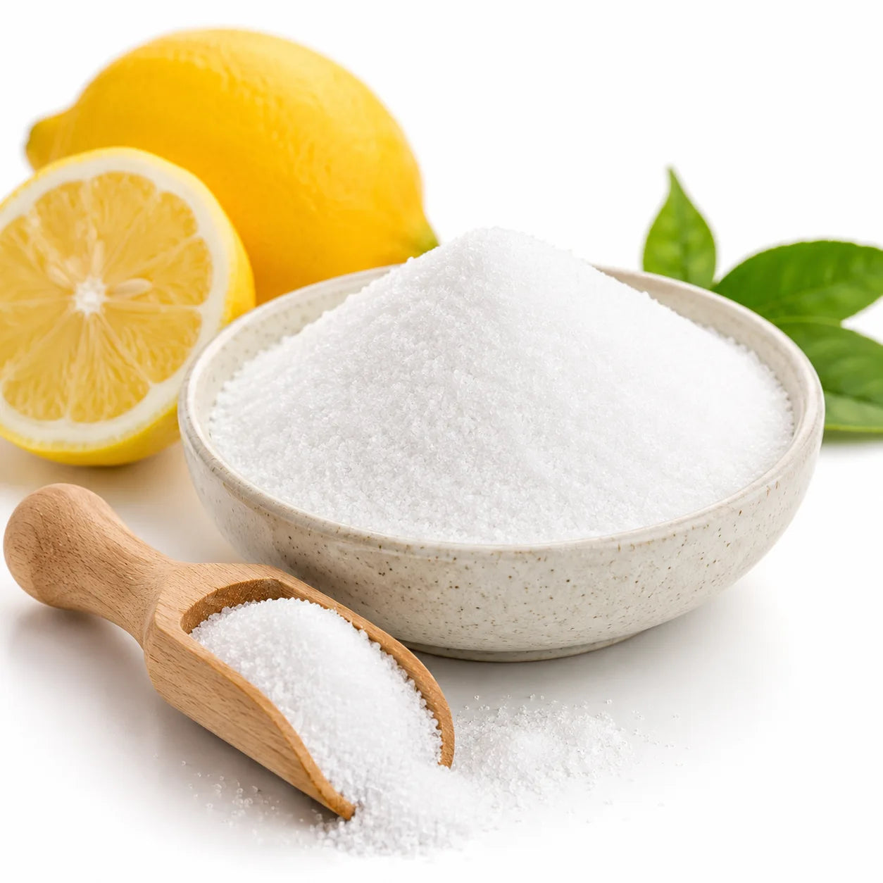 White granulated citric acid in a speckled beige ceramic bowl with a small wooden scoop in front, styled with whole and sliced lemons and green leaves on a white background.
