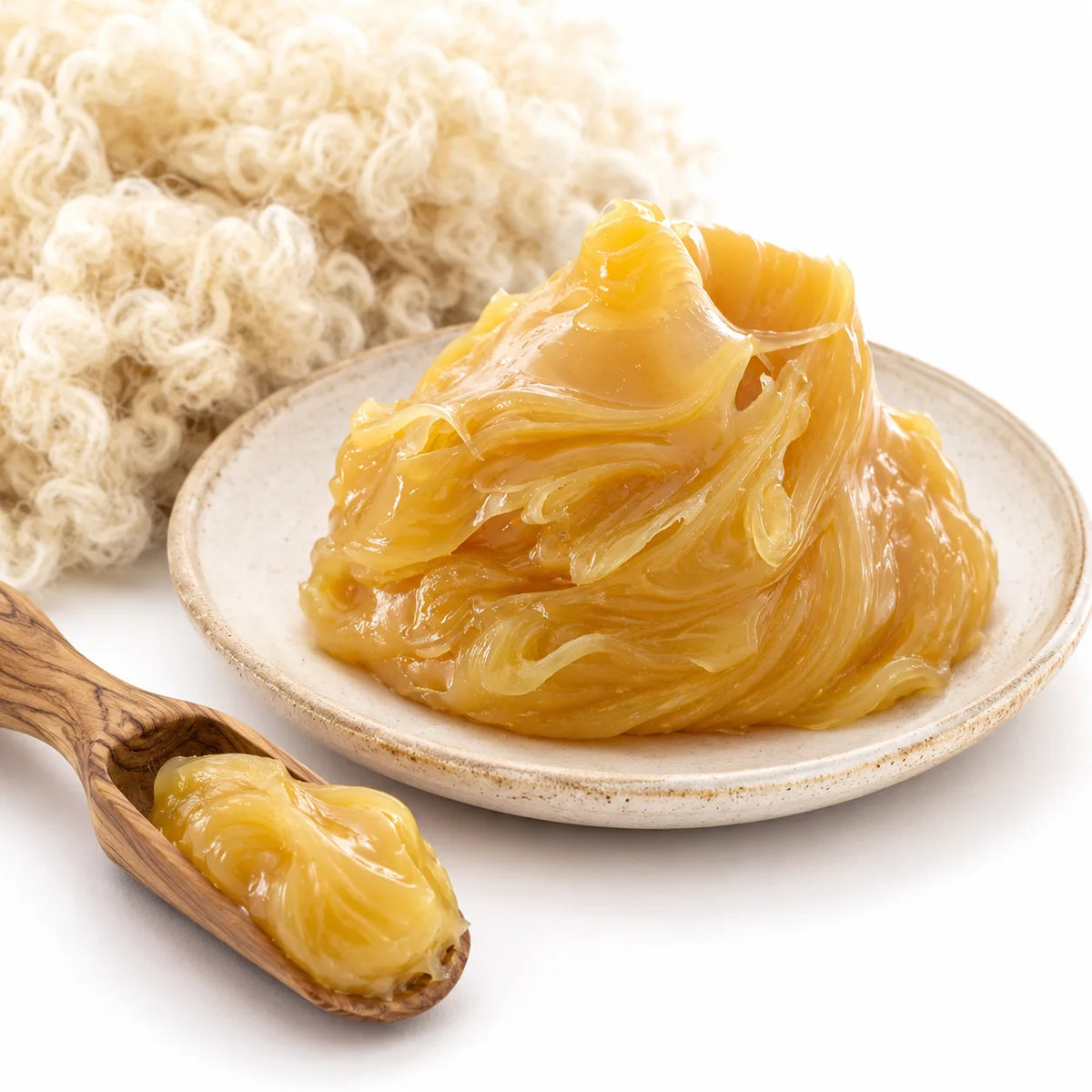 A close-up of glossy, translucent golden-yellow anhydrous lanolin piled on a small beige plate, with a wooden spoon holding some of the substance in the foreground and a bundle of off-white curly fibers behind it on a white background.
