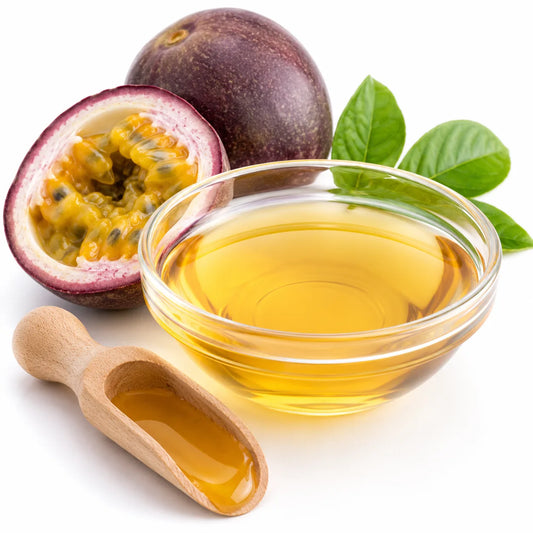 A glass bowl filled with golden passionfruit flavour oil, shown with a halved passion fruit, a whole passion fruit, green leaves, and a small wooden spoon on a white background.